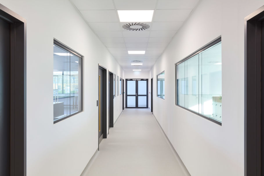 A clean, white corridor extends with closed black doors and windows lining the walls, under fluorescent ceiling lights, leading to double glass doors at the end.