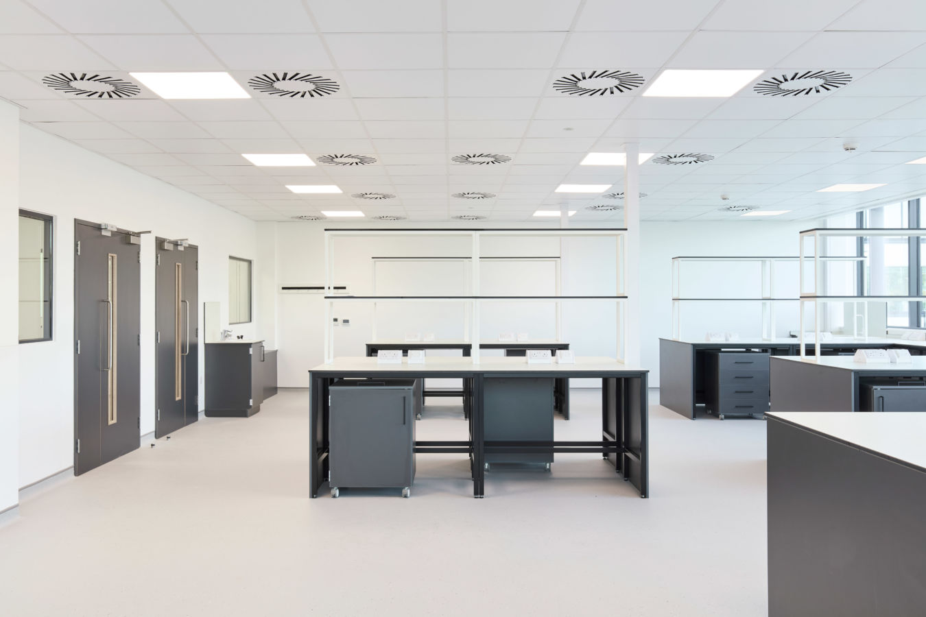 Spacious, modern laboratory with clean white walls, large central workstations, black cabinetry, and ceiling-mounted ventilation units.