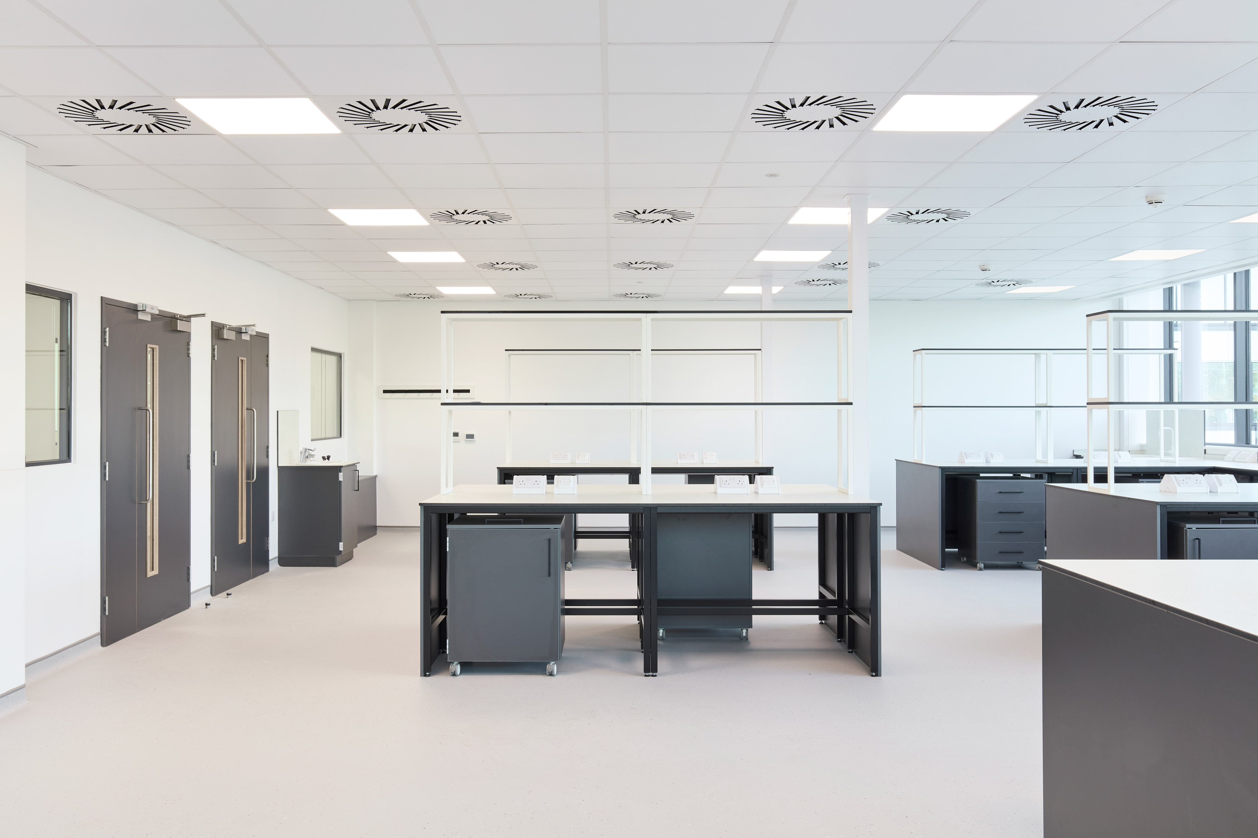 Spacious, modern laboratory with clean white walls, large central workstations, black cabinetry, and ceiling-mounted ventilation units.