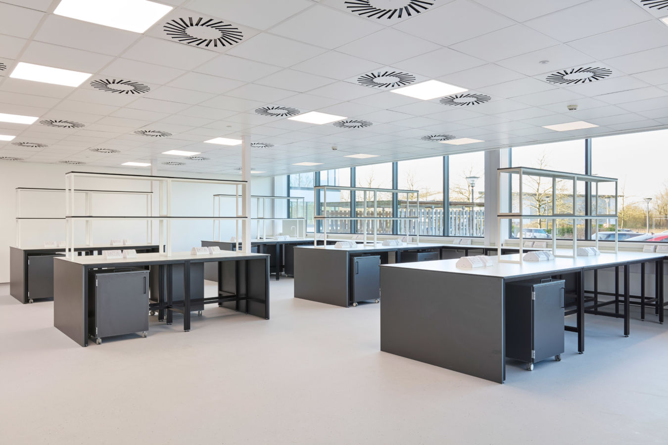 Bright and spacious laboratory with black workbenches, white countertops, large windows providing natural light, and circular ceiling-mounted ventilation units.