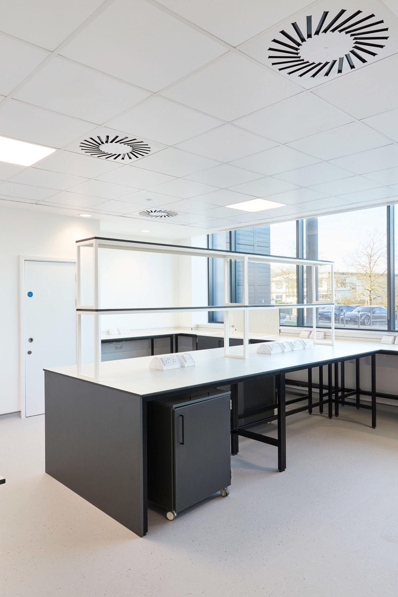Bright and spacious laboratory with black workbenches, white countertops, large windows providing natural light, and ceiling-mounted ventilation units.