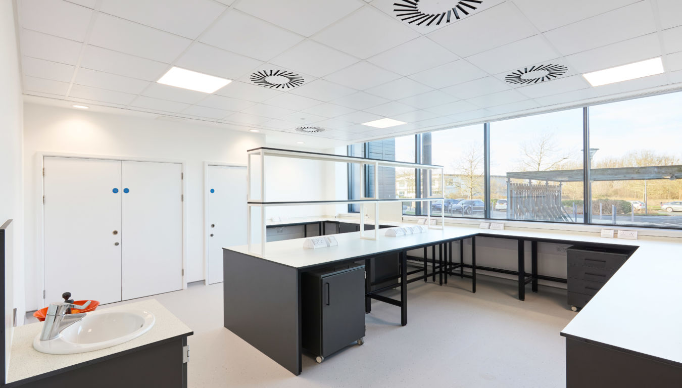 Bright and spacious laboratory with black workbenches, white countertops, large windows providing natural light, and ceiling-mounted ventilation units.