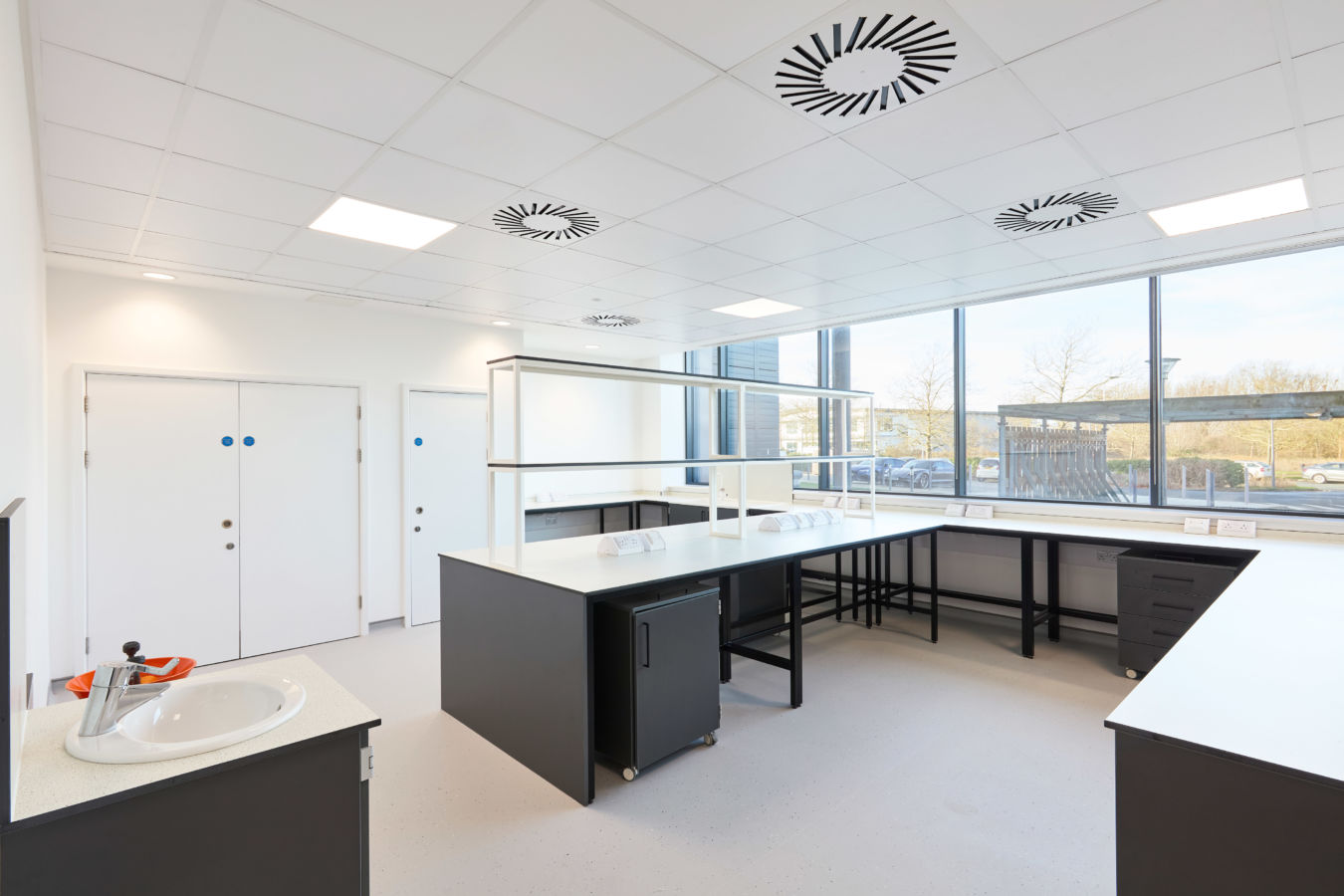 Bright and spacious laboratory with black workbenches, white countertops, large windows providing natural light, and ceiling-mounted ventilation units.