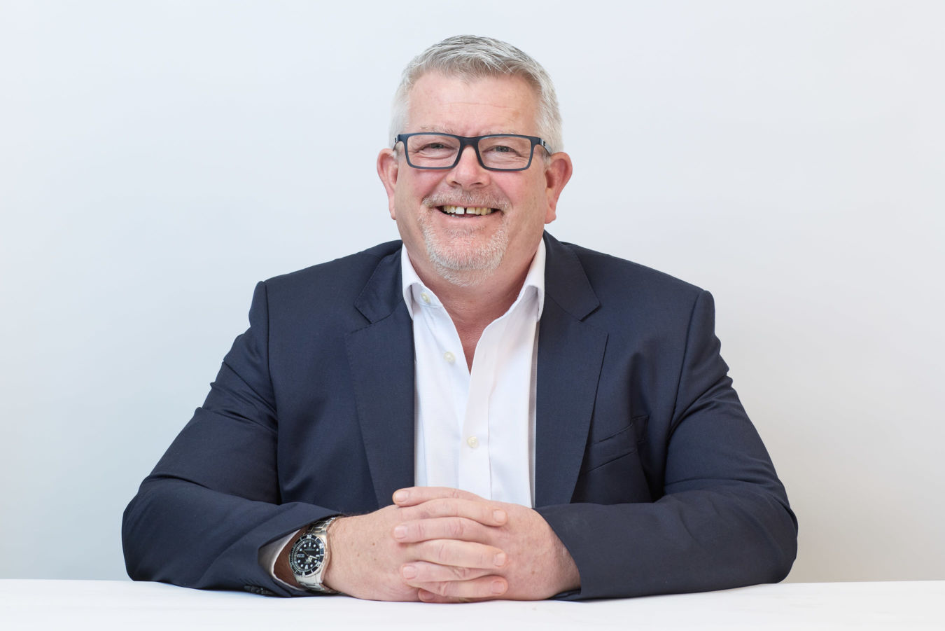 Smiling man in a dark blazer and white shirt, seated with hands clasped on a white table, wearing glasses and a wristwatch, against a plain white background.