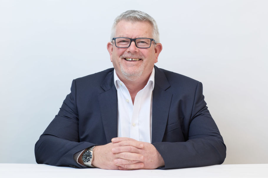 Smiling man in a dark blazer and white shirt, seated with hands clasped on a white table, wearing glasses and a wristwatch, against a plain white background.