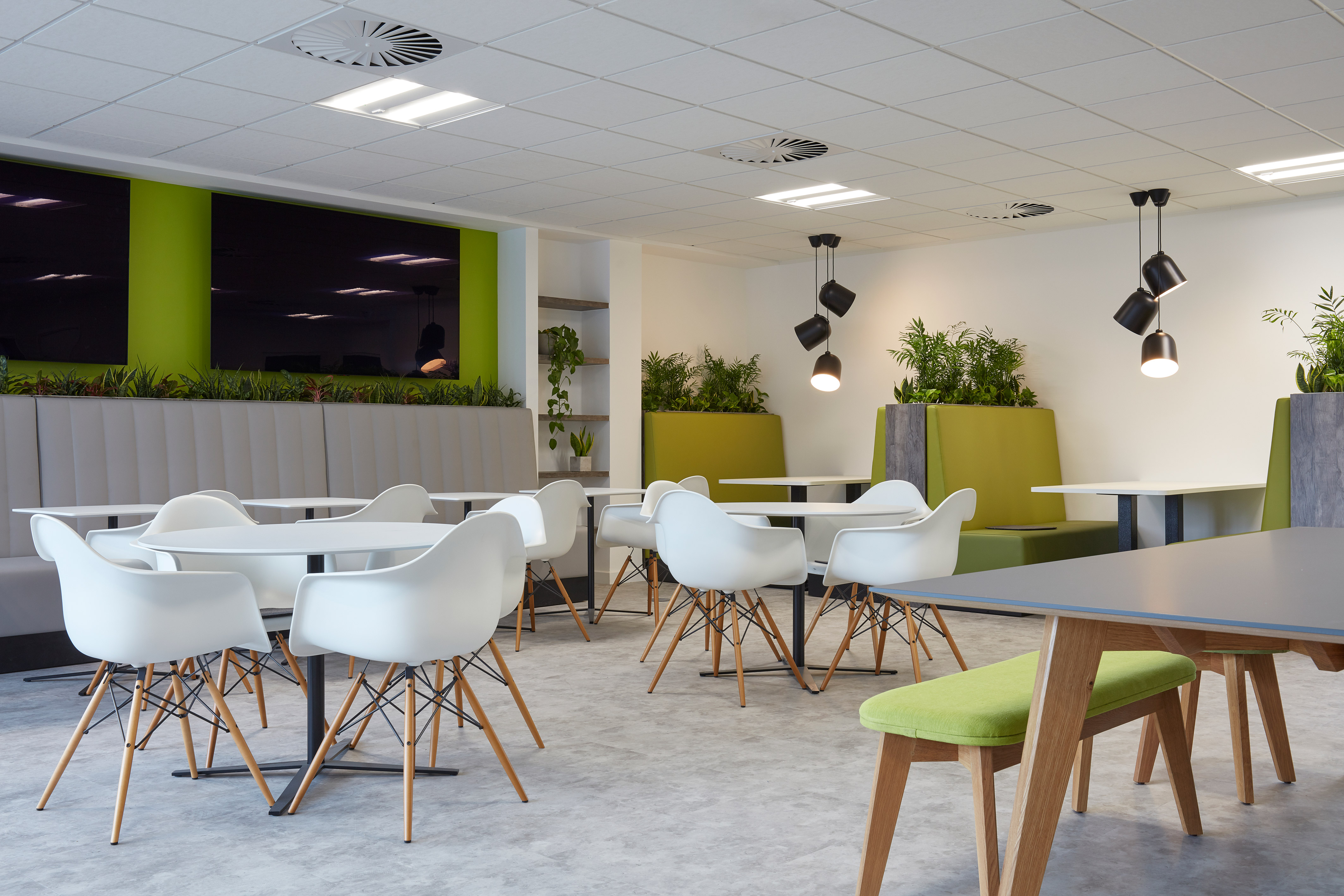 Modern cafeteria featuring white chairs around round tables; planters with greenery, pendant lights, and a TV enhance the sleek, open interior with green and gray accents.