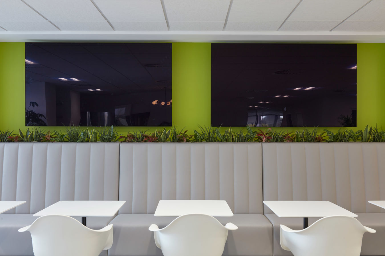 Modern seating area featuring white tables and chairs in front of a gray, cushioned bench. Green wall with plants separates two large, reflective screens above.