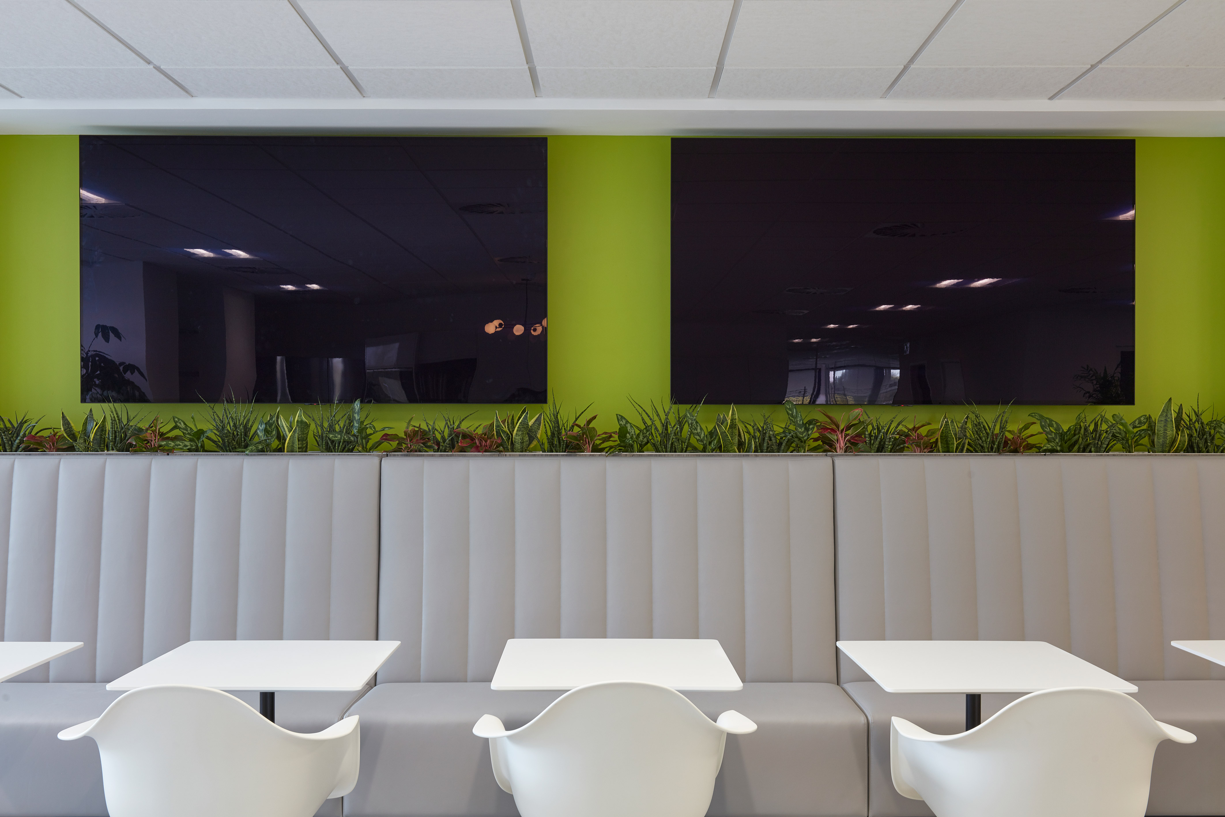 Modern seating area featuring white tables and chairs in front of a gray, cushioned bench. Green wall with plants separates two large, reflective screens above.