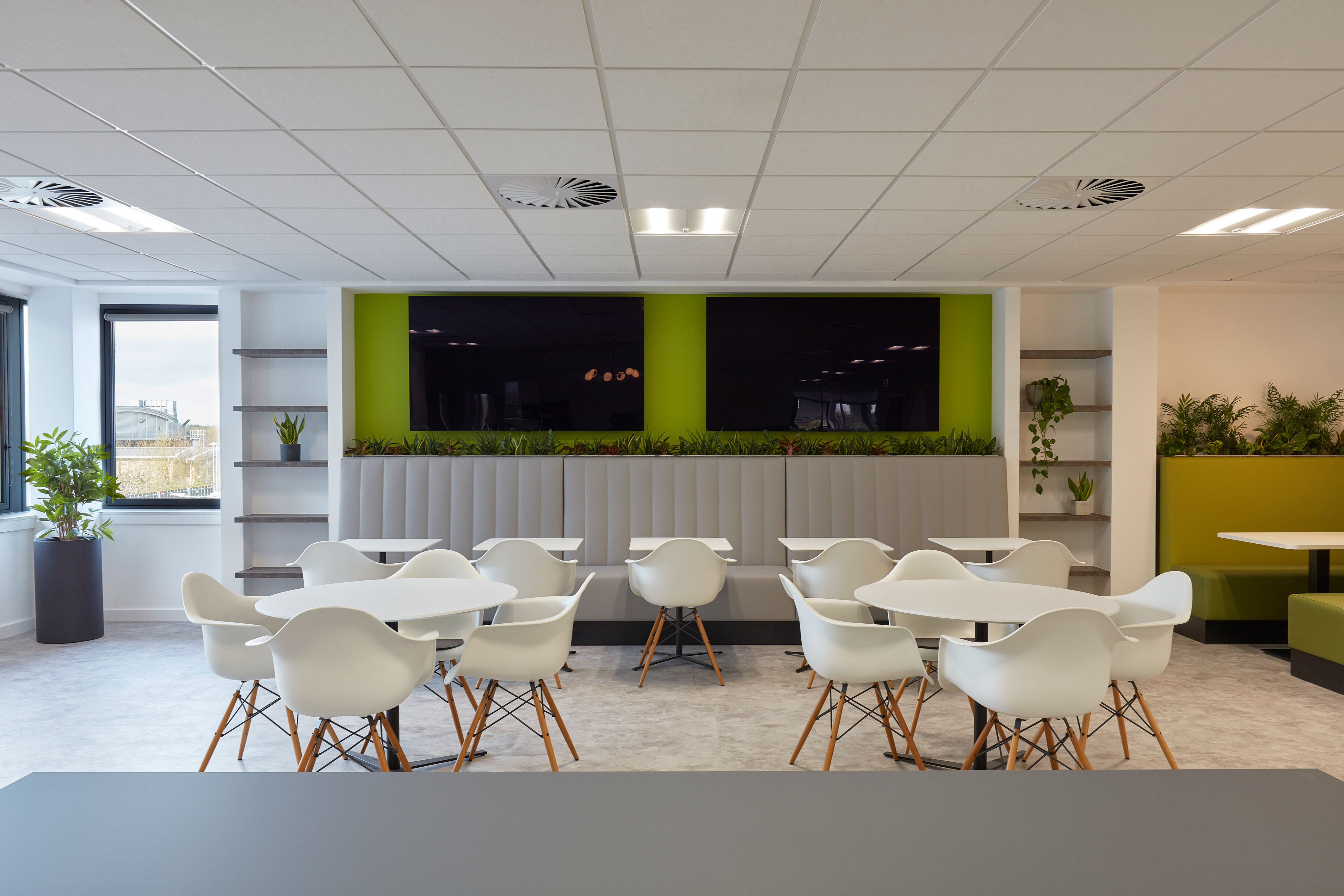 White chairs and round tables arranged in a modern office break room with green accents and plants, flanked by large windows and shelves displaying potted plants.