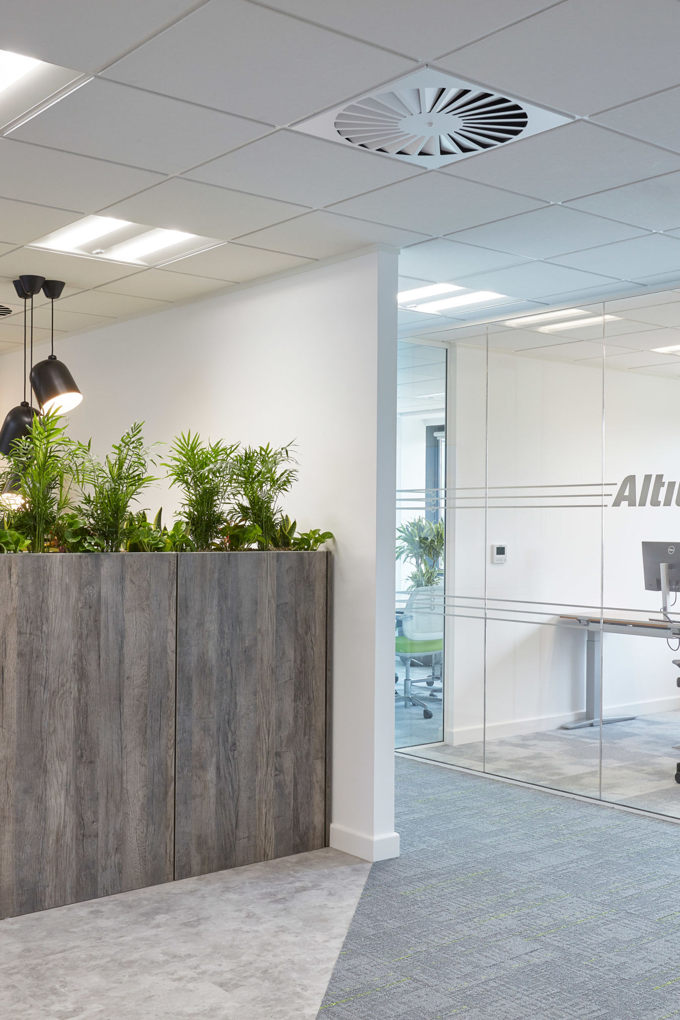 Office interior with a wooden planter filled with greenery, adjacent to a glass-walled meeting room with the text "Altu." Ceiling features lights and an air vent.