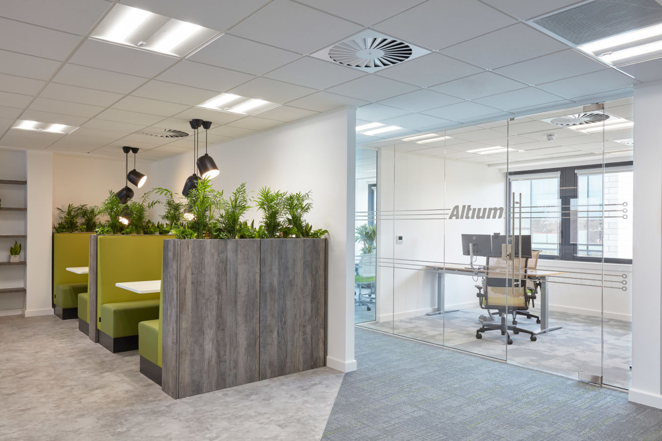 Office interior with a wooden planter filled with greenery, adjacent to a glass-walled meeting room with the text "Altu." Ceiling features lights and an air vent.