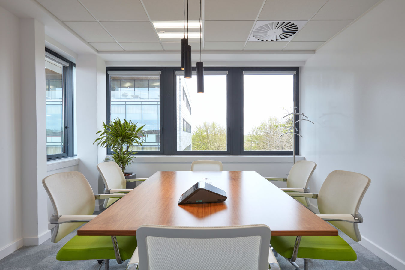 A conference room table surrounded by six chairs and a speakerphone, with large windows providing natural light and views of neighboring buildings and greenery, featuring modern ceiling lights.