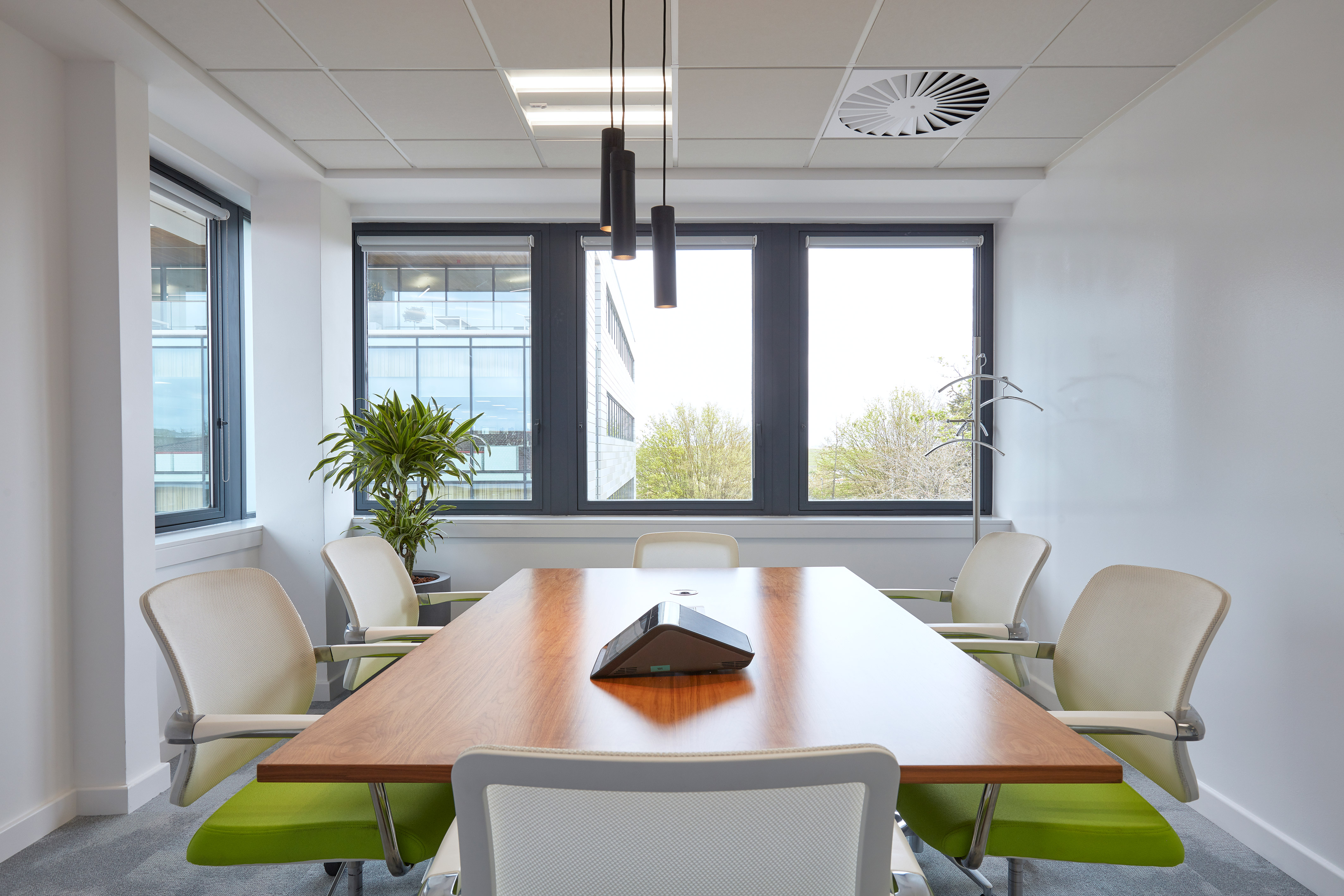 A conference room table surrounded by six chairs and a speakerphone, with large windows providing natural light and views of neighboring buildings and greenery, featuring modern ceiling lights.