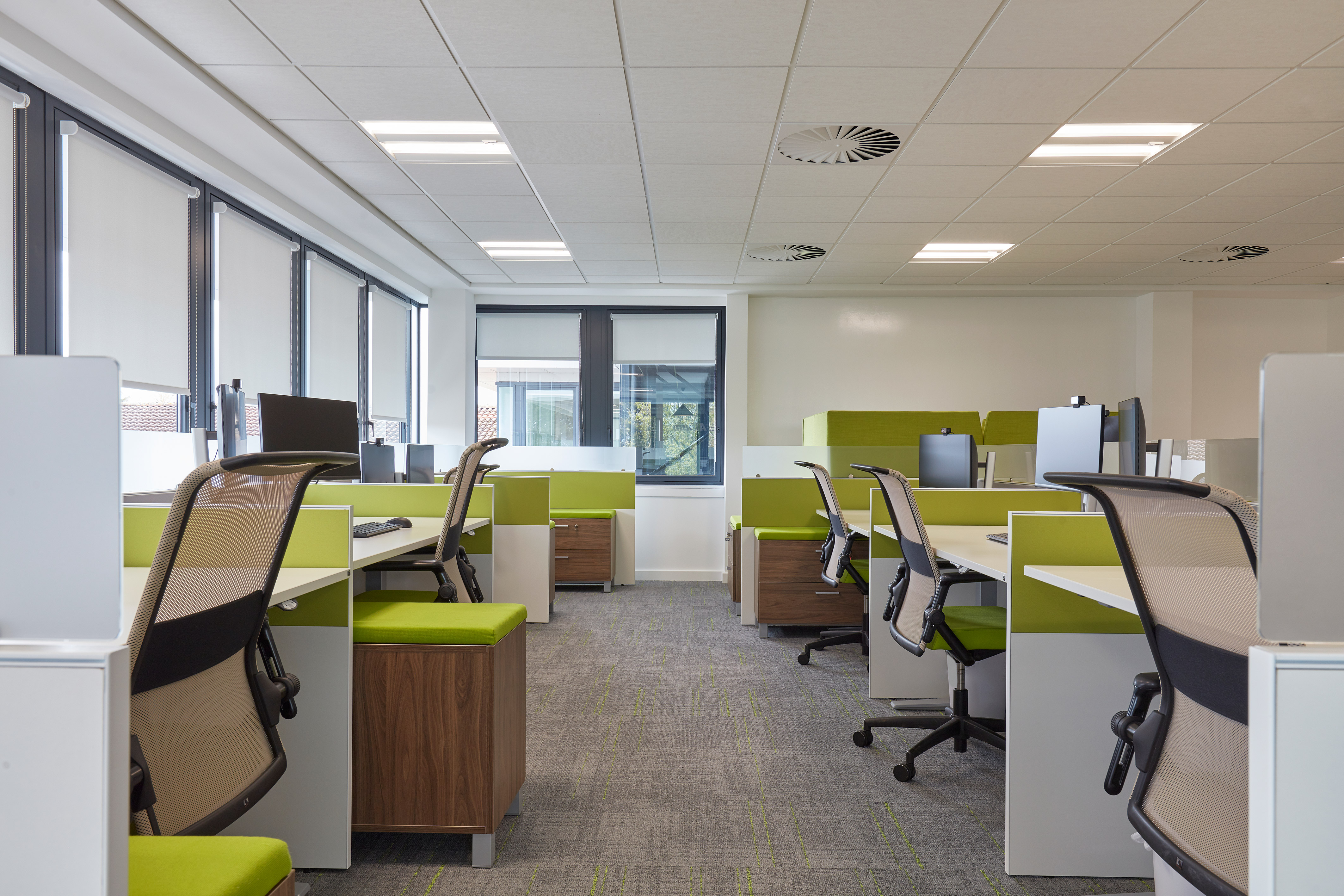 Office desks with green chairs and computer monitors are arranged in neat rows, surrounded by large windows with blinds, under a suspended ceiling with lighting and ventilation panels.