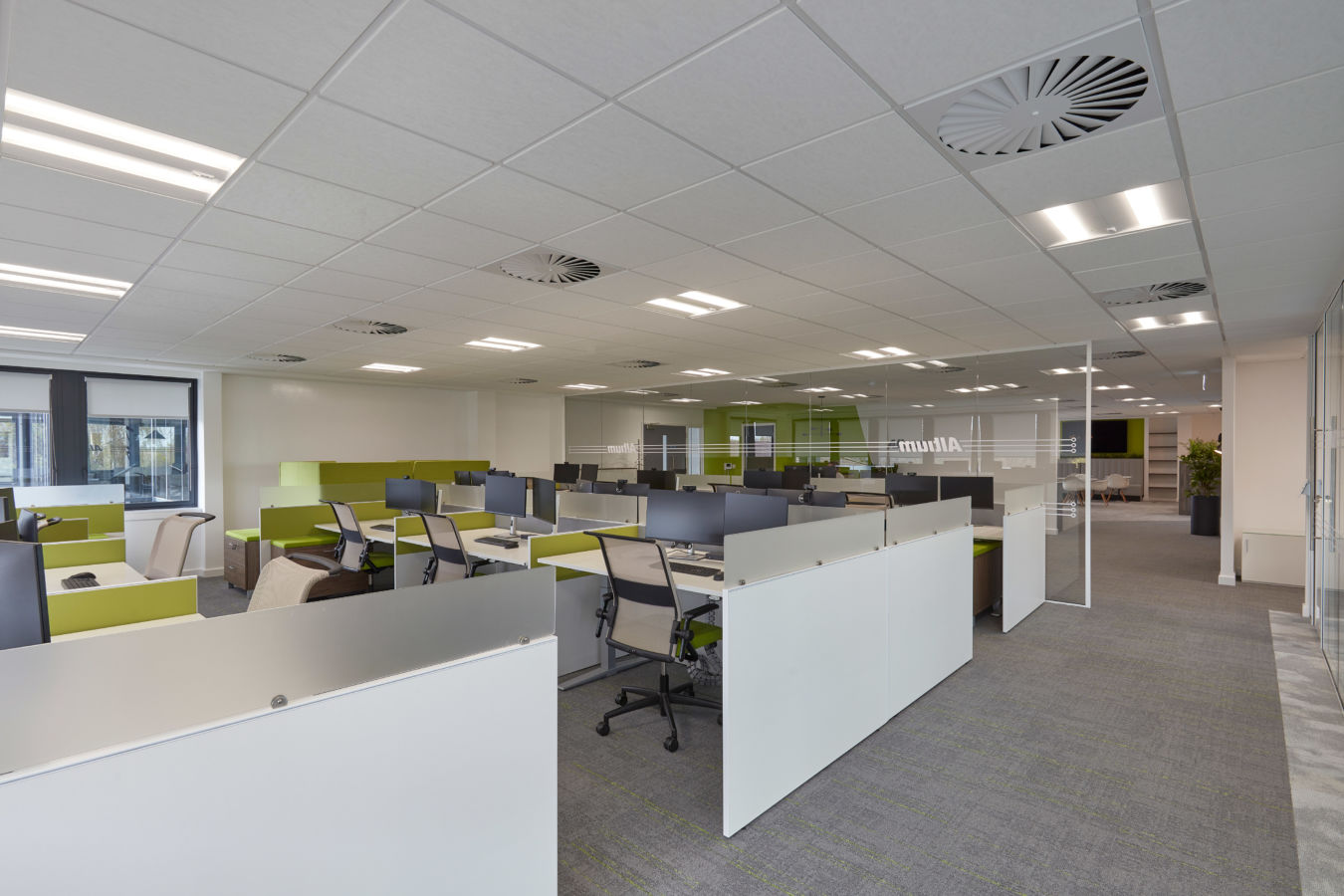 Open-plan office with green and white workstations, ergonomic chairs, and computers, in a spacious area with ceiling lights, glass dividers, and the word "Altium" visible on a partition.