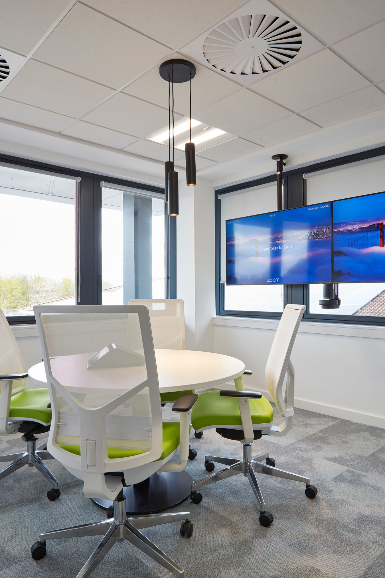 Modern conference room with a round table, four white chairs with green cushions, and dual screens on the wall showing a cityscape. Bright with large windows