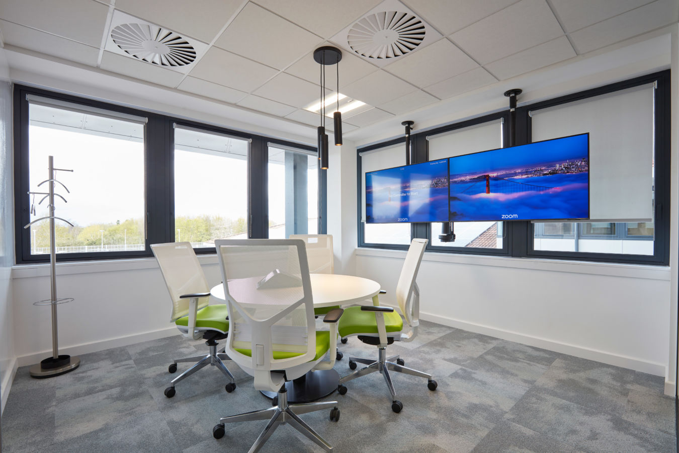 Modern conference room with a round table, four white chairs with green cushions, and dual screens on the wall showing a cityscape. Bright with large windows