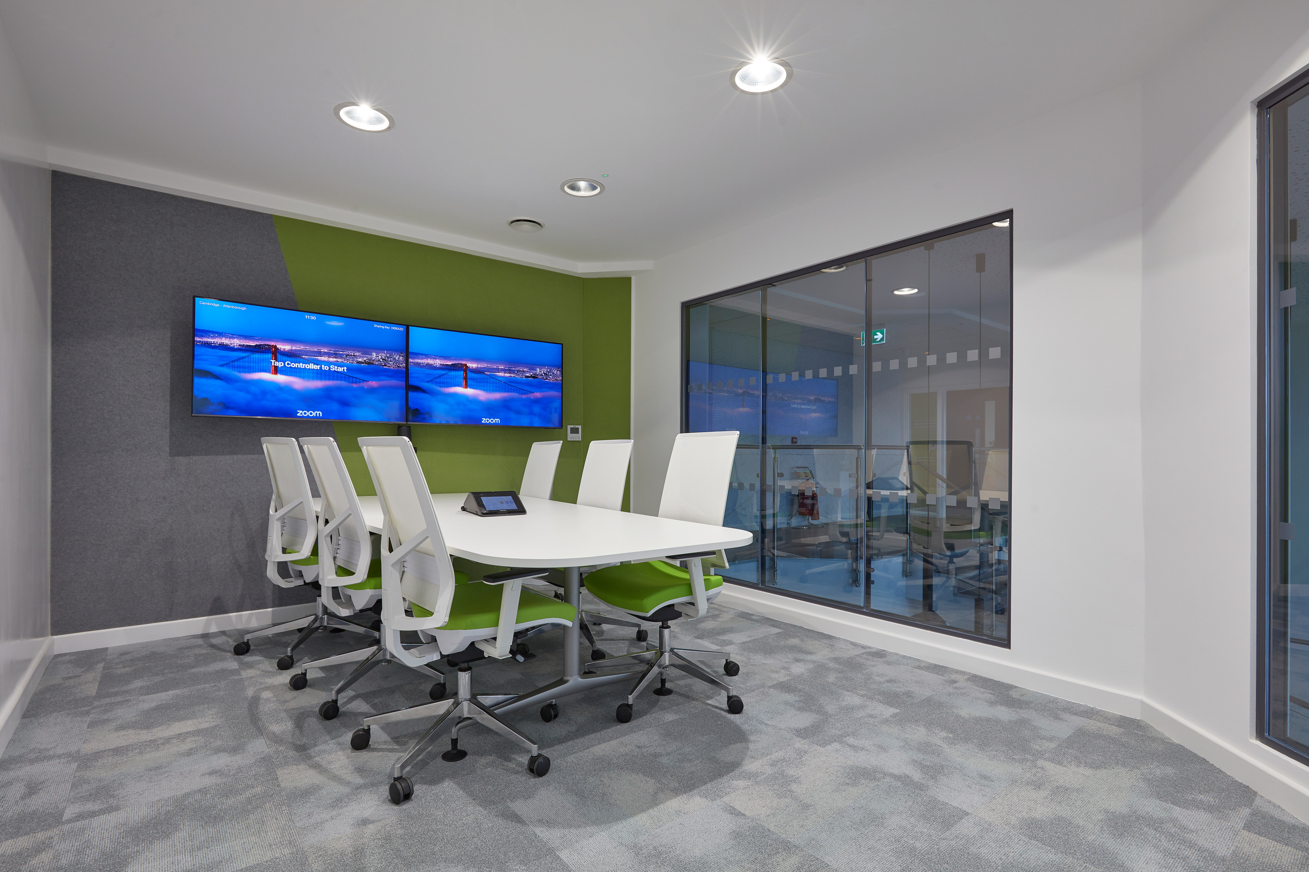 Conference room featuring a white oval table with six white chairs on wheels, facing two wall-mounted screens displaying "Zoom." A large interior glass window separates the space.
