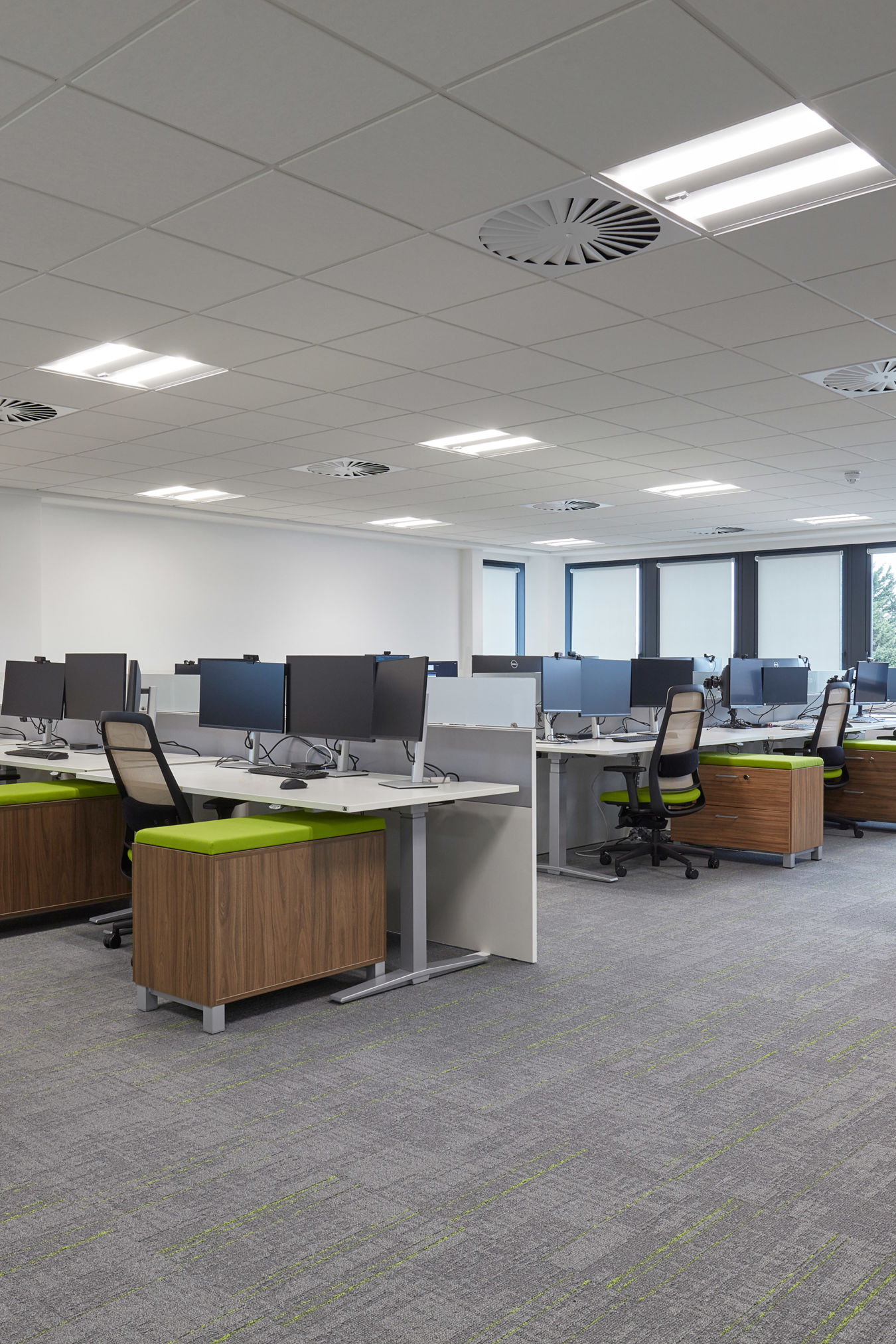 A modern office space with rows of desks featuring dual monitors and swivel chairs. Green accents on chairs and natural light through large windows create a fresh, inviting atmosphere.