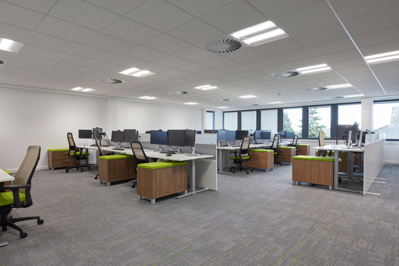 A modern office space with rows of desks featuring dual monitors and swivel chairs. Green accents on chairs and natural light through large windows create a fresh, inviting atmosphere.