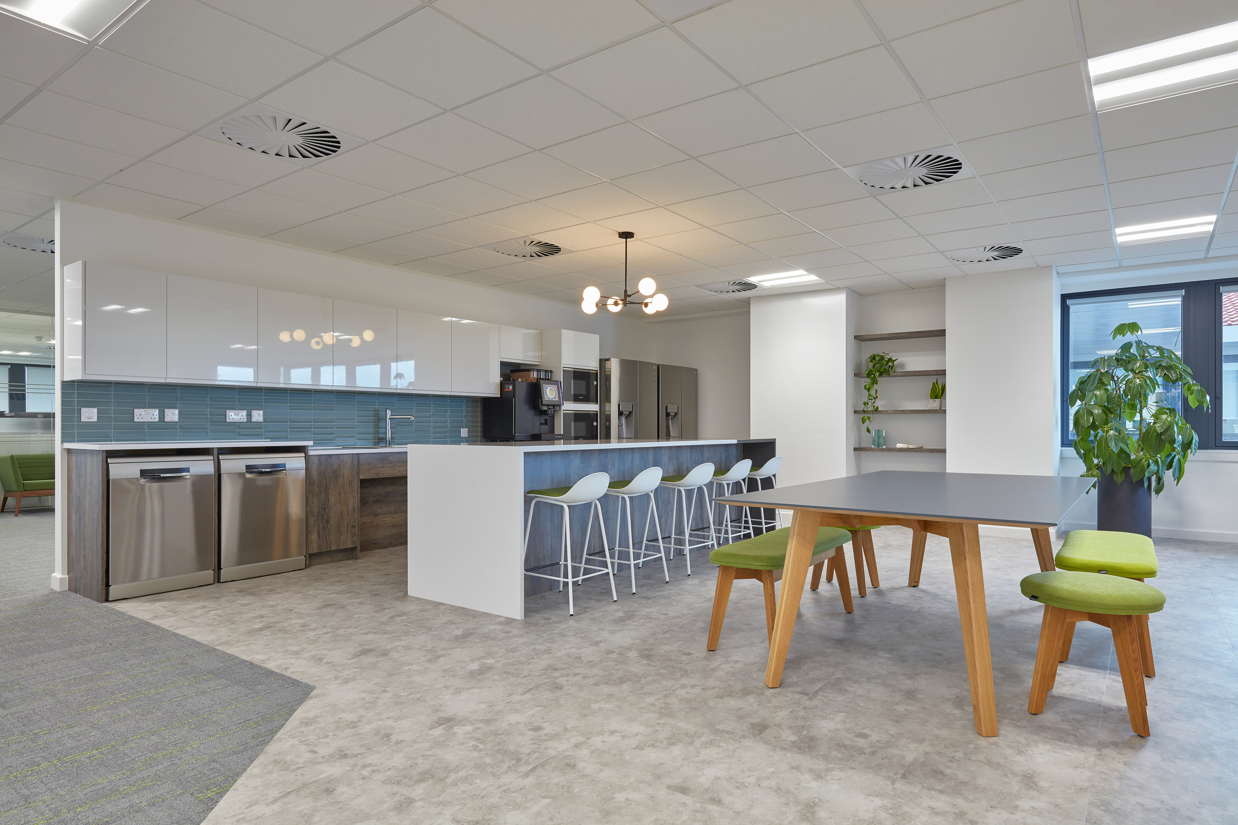 Modern kitchen area with a long counter and white bar stools, surrounded by sleek cabinetry. A dining table with green seats and a large plant complete the spacious, minimalistic setting.