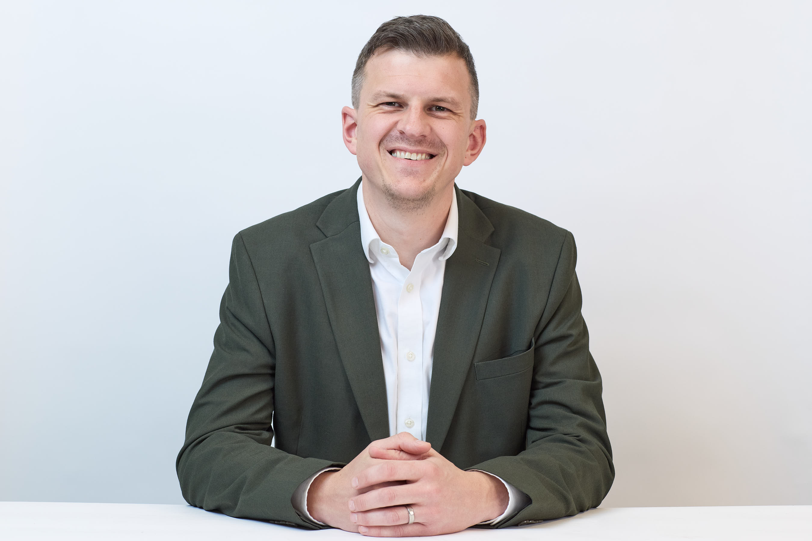 A person in a green suit and white shirt smiles while sitting at a white table against a plain white background.