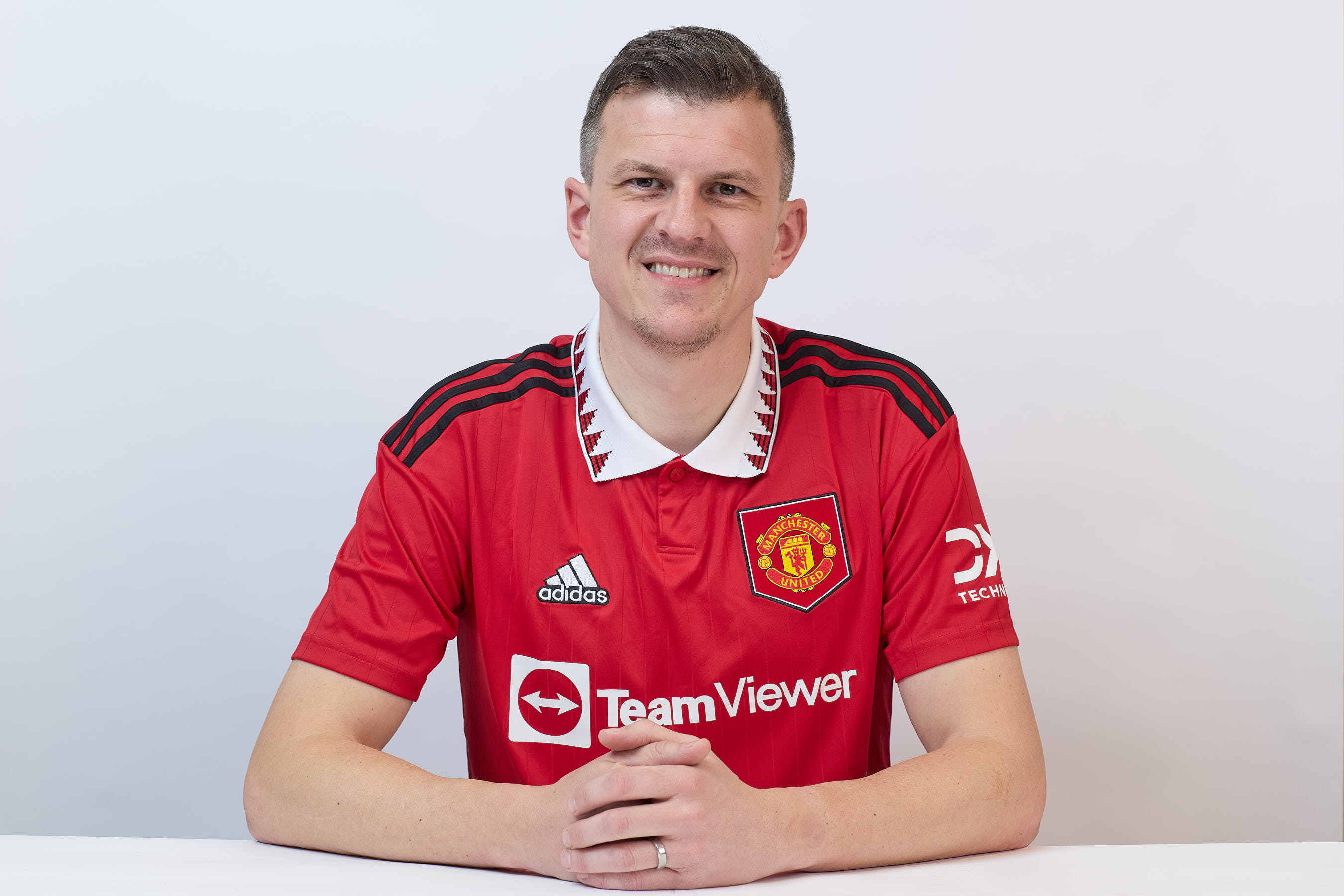 A person wearing a red Manchester United jersey with "adidas," "TeamViewer," and "DXC Technology" logos sits with hands clasped against a white background, smiling at the camera.