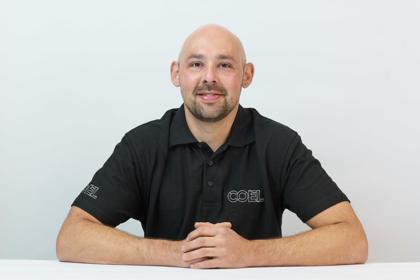A person wearing a black polo shirt with the logo "COEL" sits at a table, hands folded, against a plain white background.