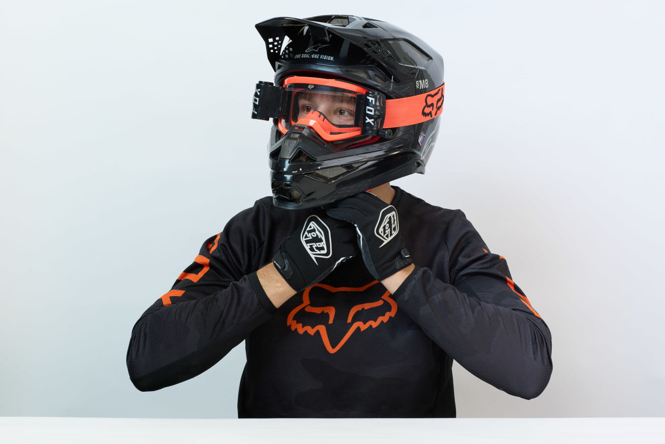 Motorcyclist wearing a black and orange helmet and gloves, adjusting gear. The helmet features the text “SM8” and “ONE GOAL. ONE VISION.” against a plain background.