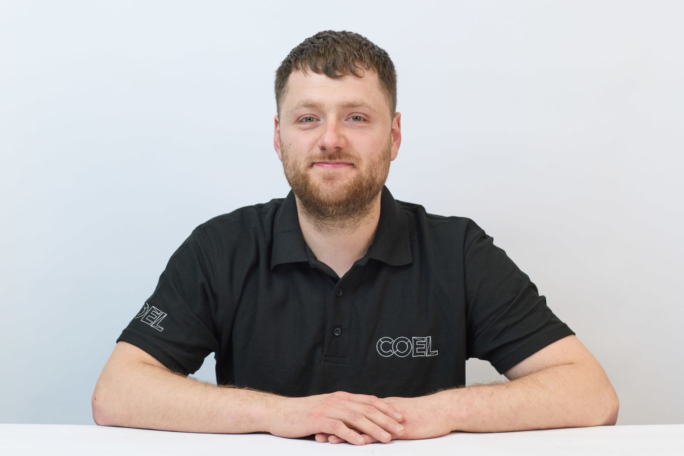 A person wearing a black polo shirt with "COEL" text, seated at a white table, against a plain white background.