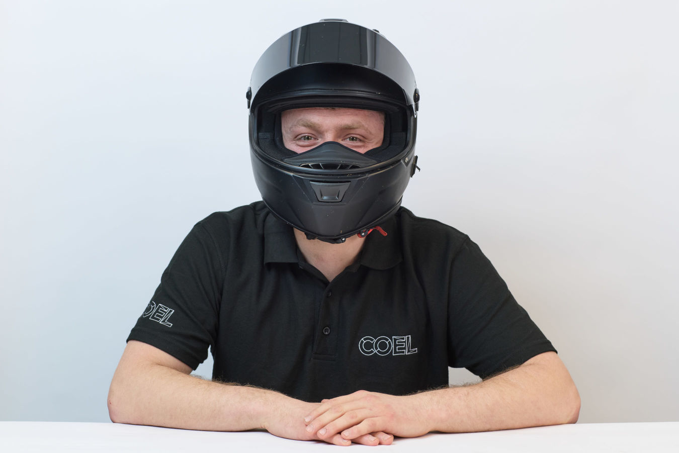 A person wearing a black motorcycle helmet and a black shirt with "COEL" embroidered on it sits against a plain white background.