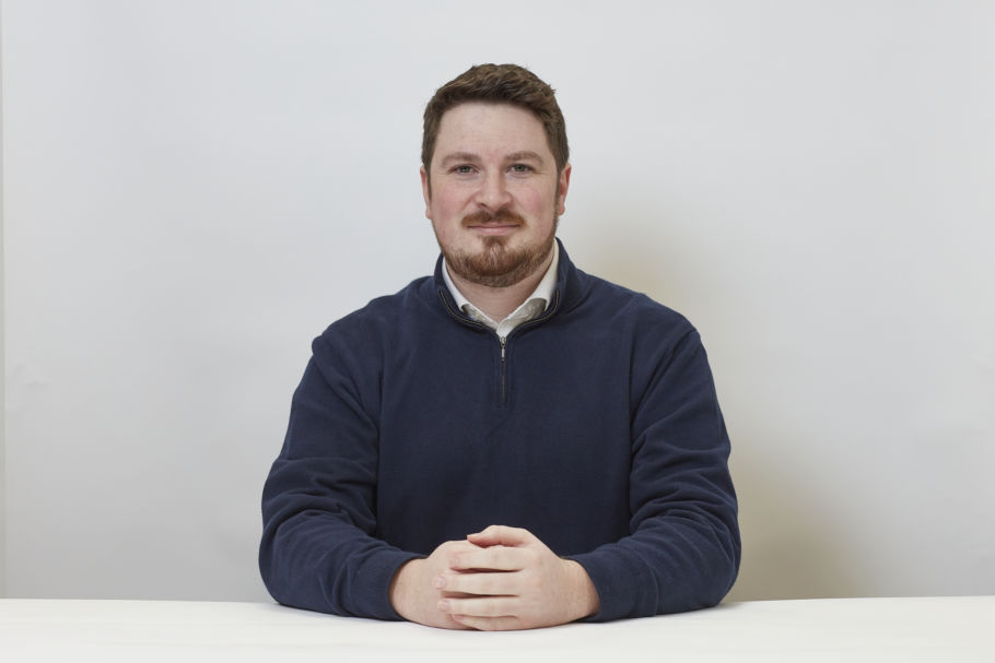 A man with short hair and a beard sits at a table, wearing a navy sweater, hands clasped, against a plain white background.