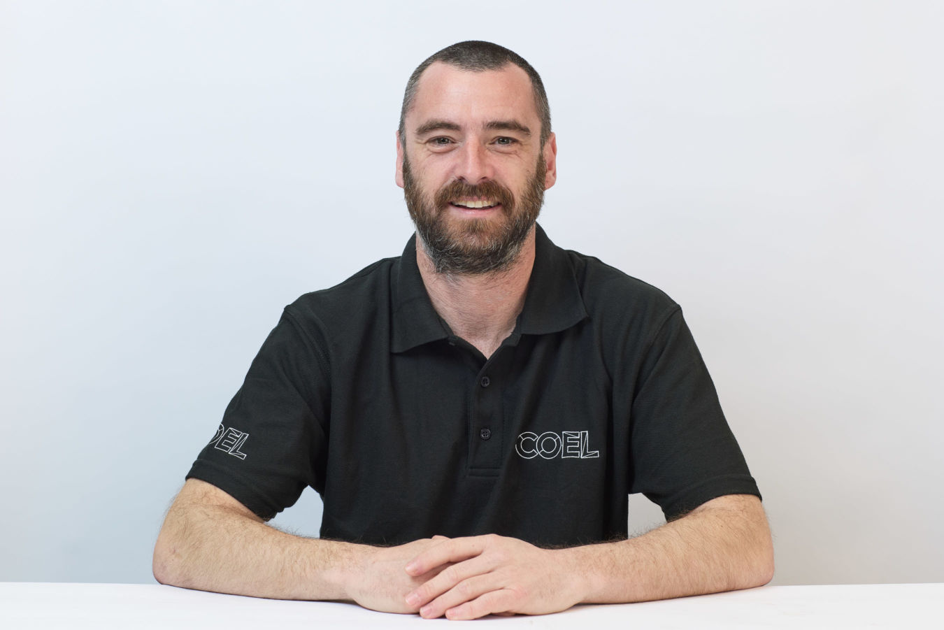 A man with a beard smiles, wearing a black polo shirt with "COEL" printed on it, sitting against a plain white background.