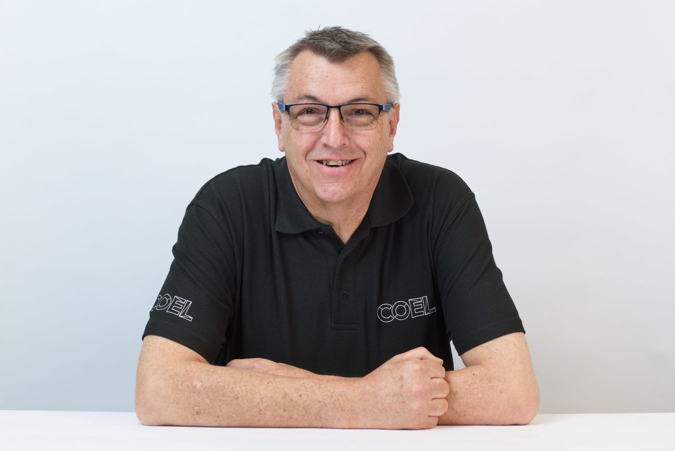 A person wearing glasses and a black polo shirt with "COEL" embroidered on the chest and sleeve is smiling while sitting at a white table against a plain background.