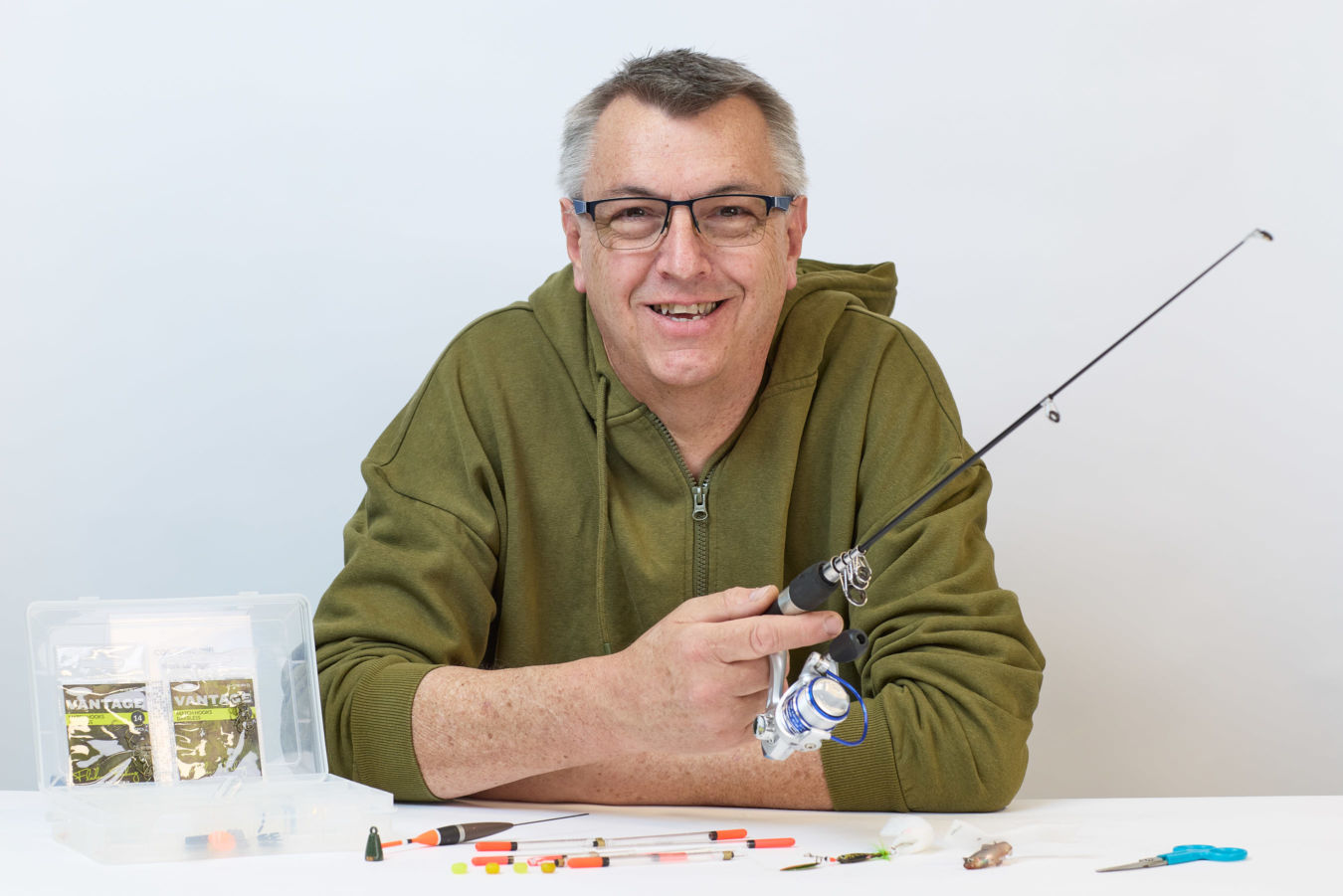 A person wearing glasses and a green hoodie holds a fishing rod, sitting at a table with fishing gear, including floats and tackle boxes labeled "Vantage," in a bright setting.
