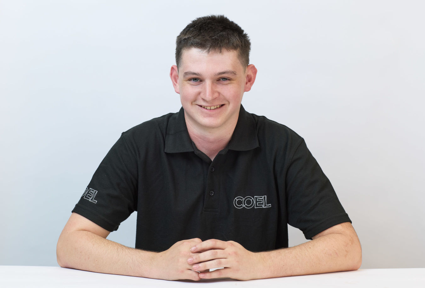 A person wearing a black polo shirt with the text "COEL" smiles while sitting at a white table against a plain background.