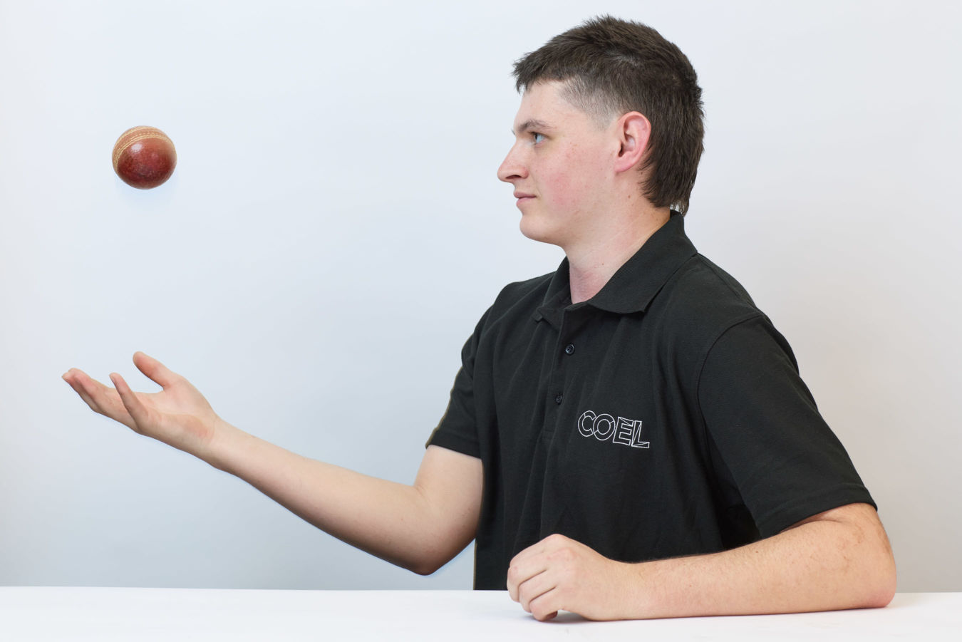 A person wearing a black shirt with "COEL" text juggles a cricket ball while seated against a plain background.