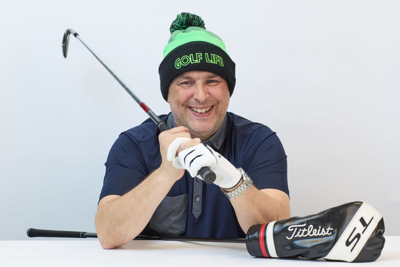 Smiling man wearing a "GOLF LIFE" beanie holds a golf club across his shoulder, with a Titleist driver headcover on a table in a bright setting.