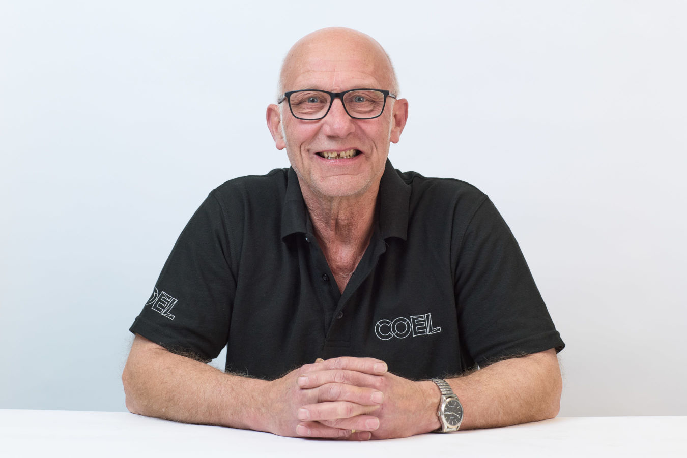 A smiling man wears glasses and a black shirt with "COEL" embroidered, sitting with hands clasped in front. He appears in a neutral, isolated white background.
