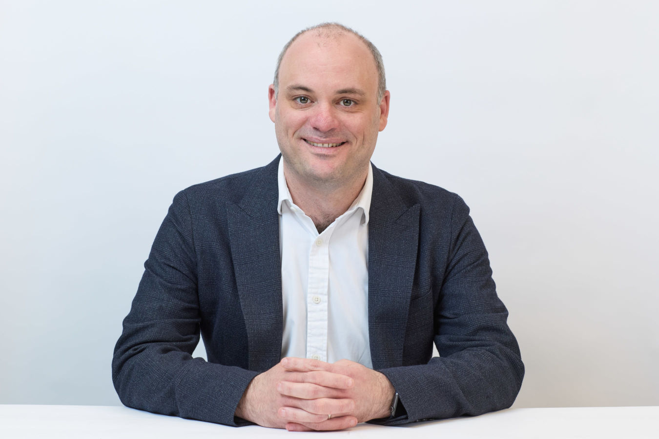 Smiling person sits at a white table with hands clasped, wearing a dark blazer and white shirt, against a plain light background.