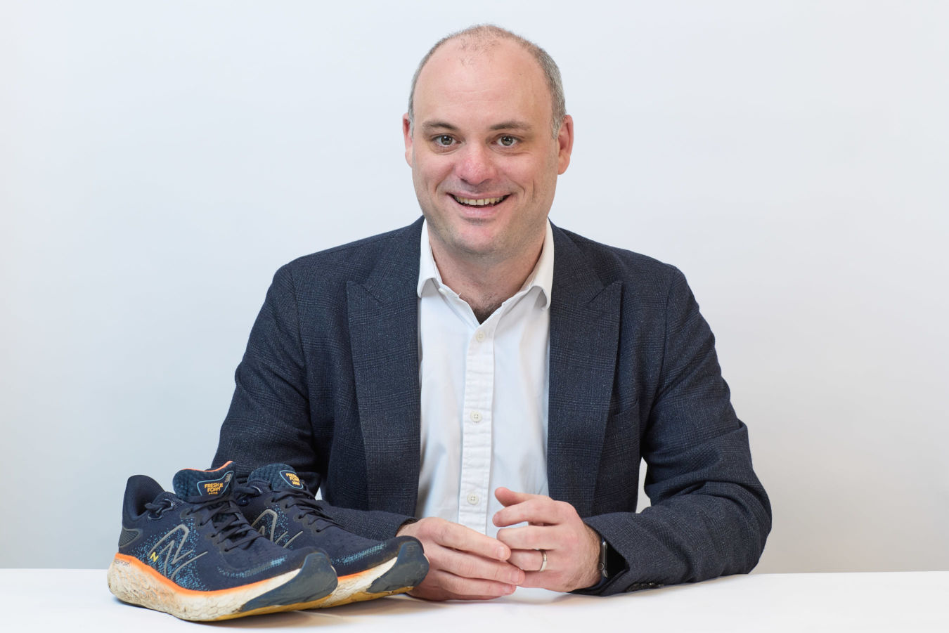 A pair of navy blue running shoes is displayed on a white table, with a smiling person in a suit sitting behind them against a plain background.