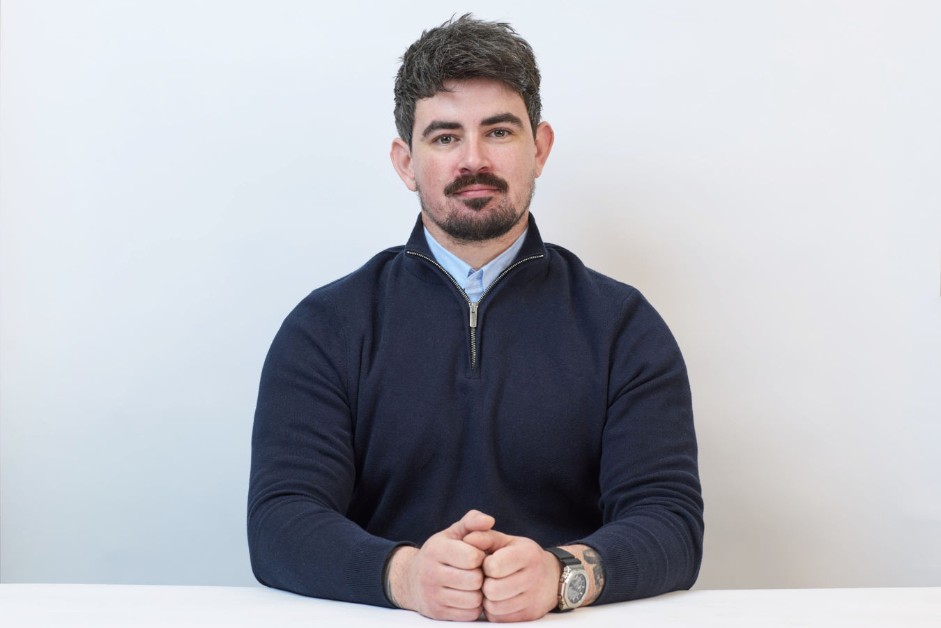 A man in a navy sweater sits with hands clasped on a white table, set against a plain light background.
