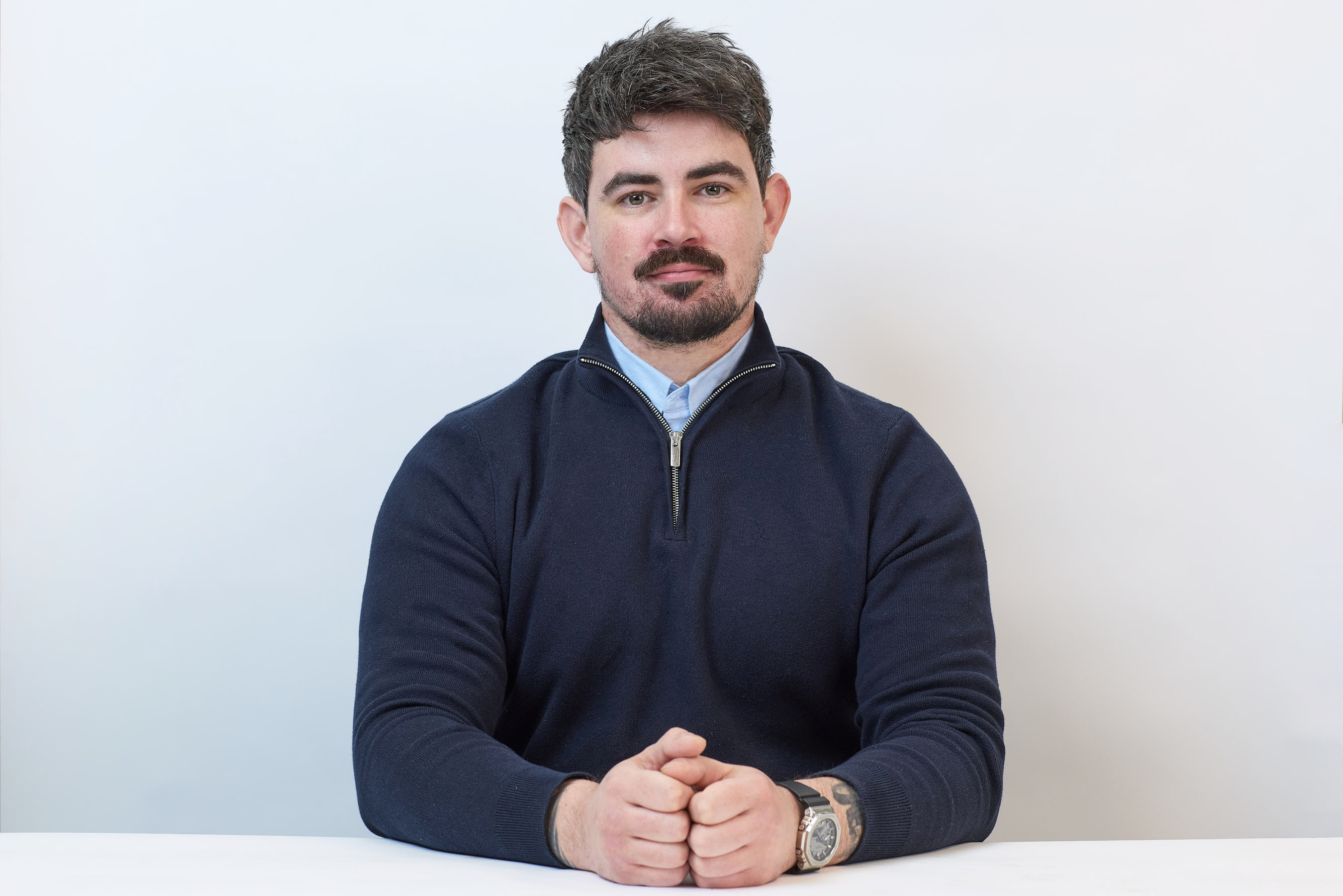 A man in a navy sweater sits with hands clasped on a white table, set against a plain light background.