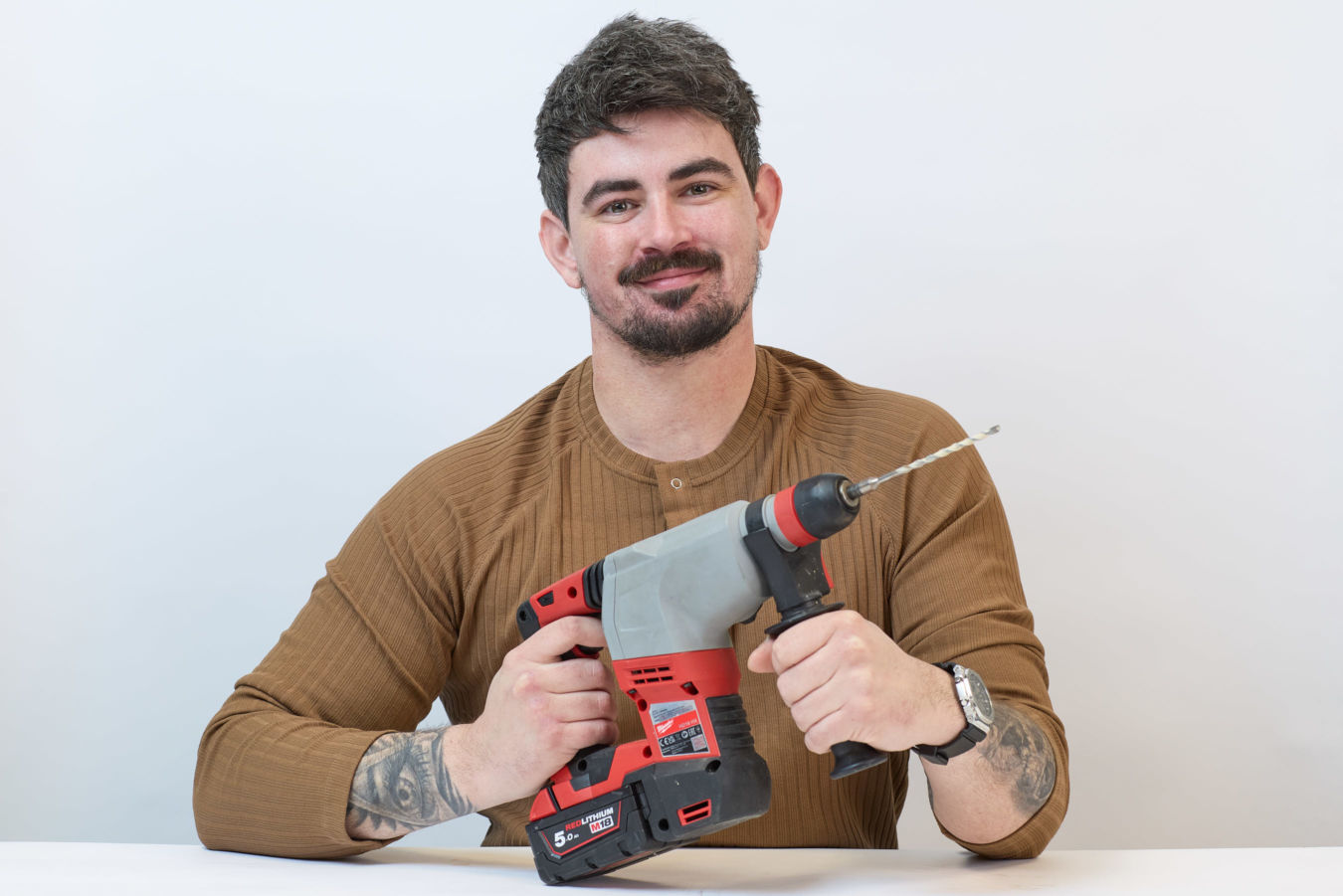 A person holds a red and gray power drill with a visible bit, sitting at a white table in a plain environment, wearing a brown top and displaying a tattoo on the arm.