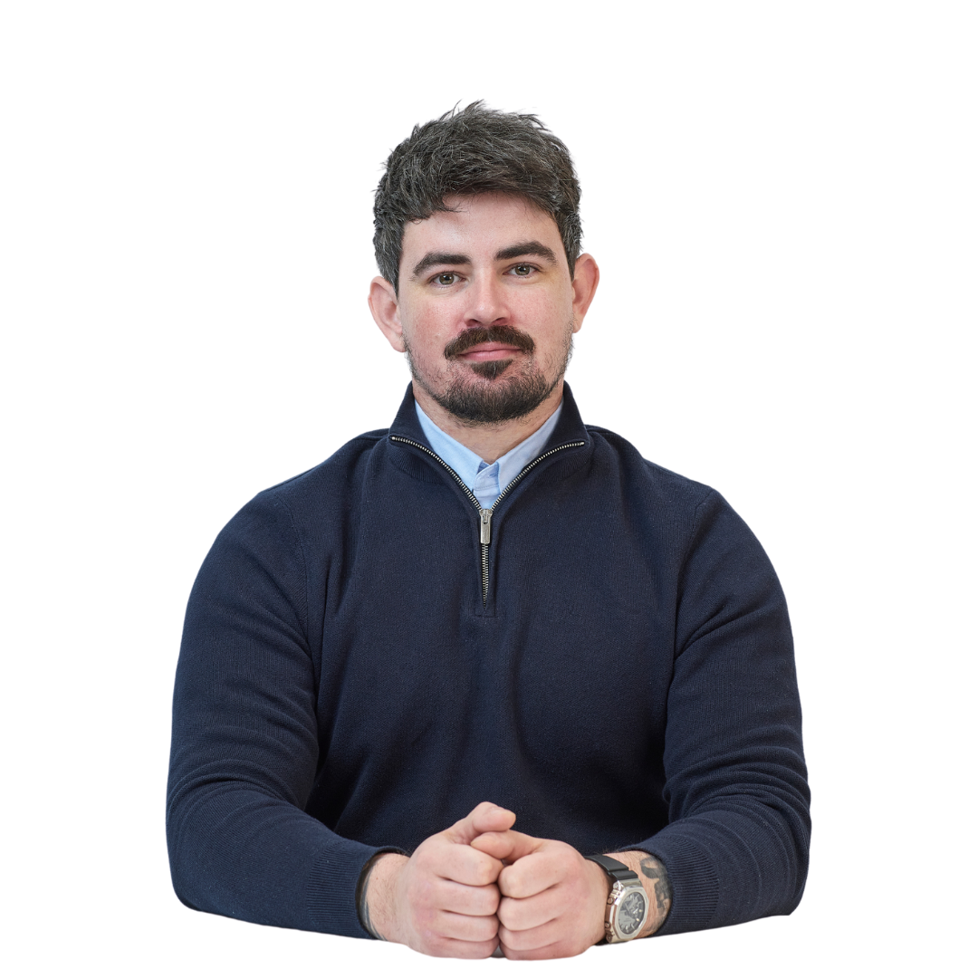 A person with short hair and a beard sits with hands clasped, wearing a dark sweater against a plain background.
