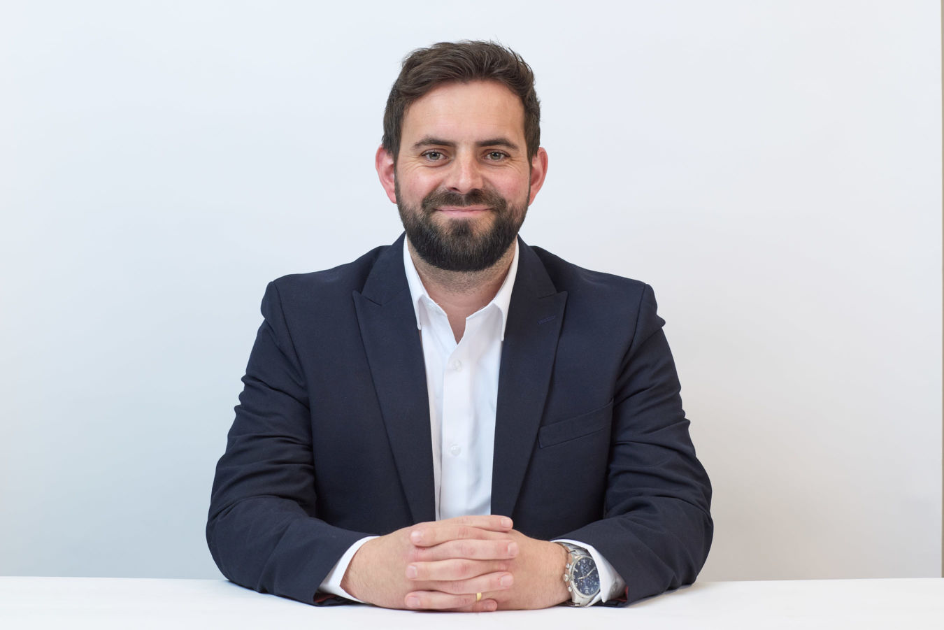 A bearded man in a navy suit sits with folded hands, smiling confidently against a white background.