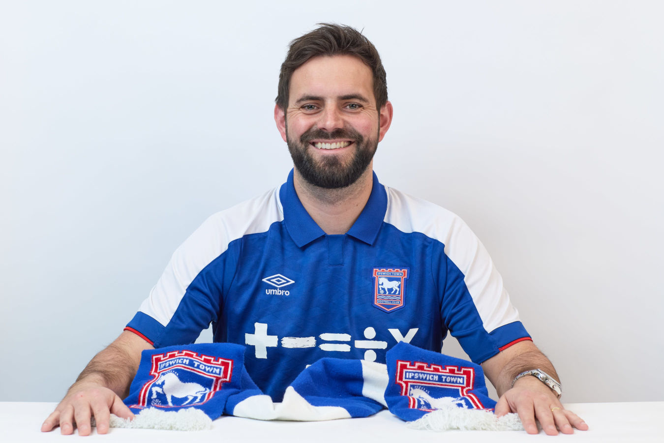A man wearing a blue Ipswich Town FC shirt smiles, sitting behind a matching scarf with the text "IPSWICH TOWN" on a white background.