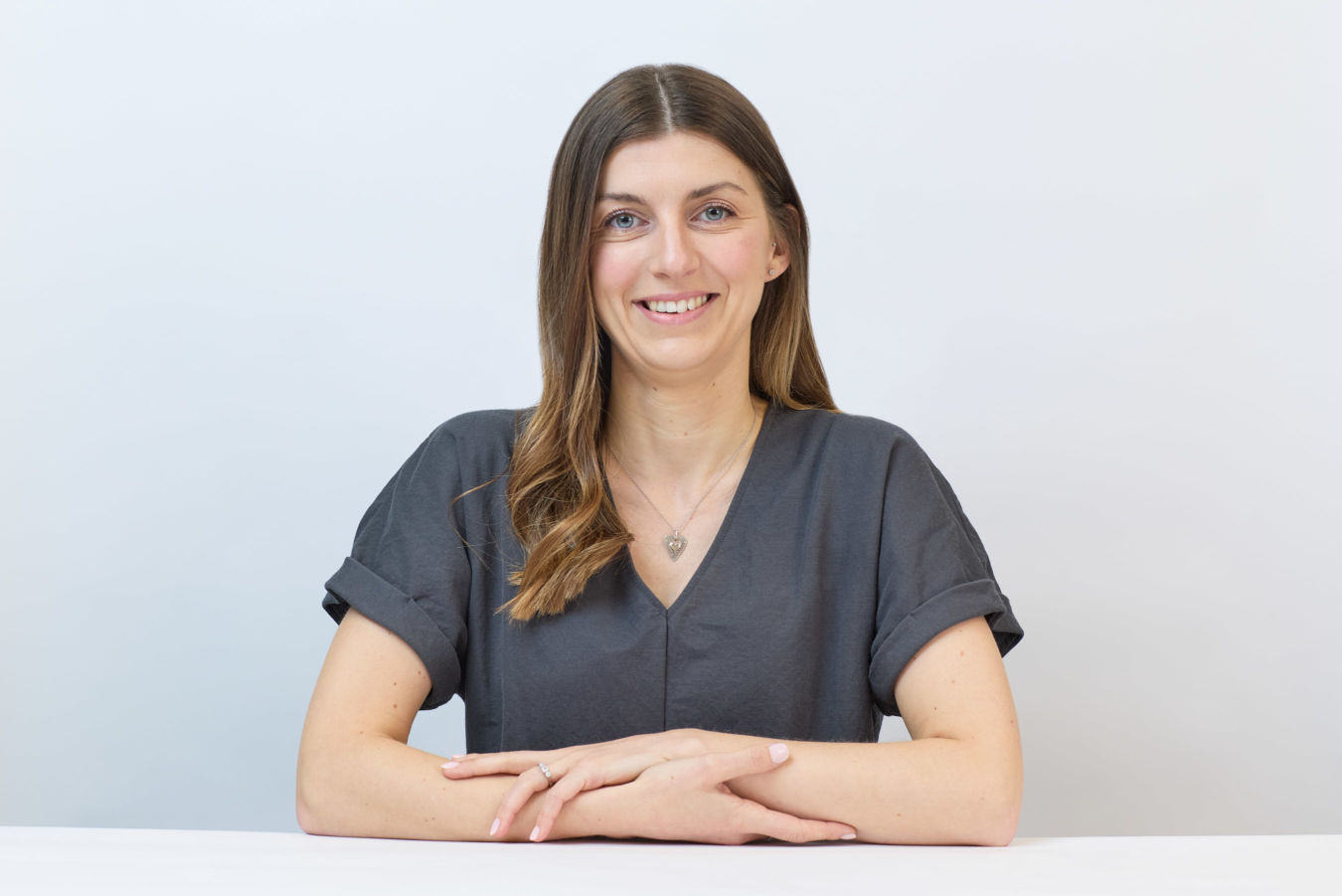 A person with long brown hair, wearing a gray shirt and heart necklace, is smiling and sitting at a white table against a plain background.