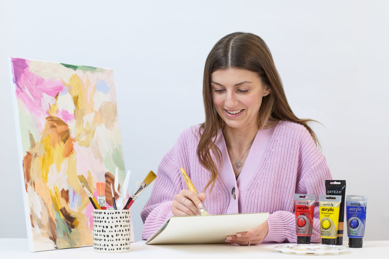 A woman in a pink sweater draws on paper at a table with acrylic paints, brushes in a holder, and an abstract painting.