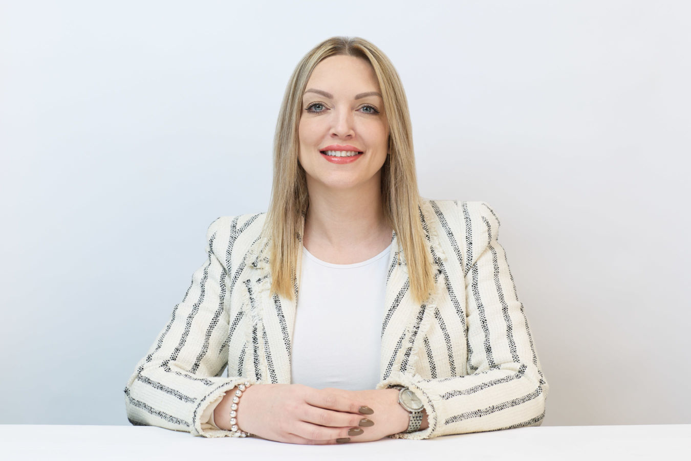 A woman with long blonde hair sits smiling, hands resting on a table. She wears a white striped blazer and a wristwatch, set against a plain white background.